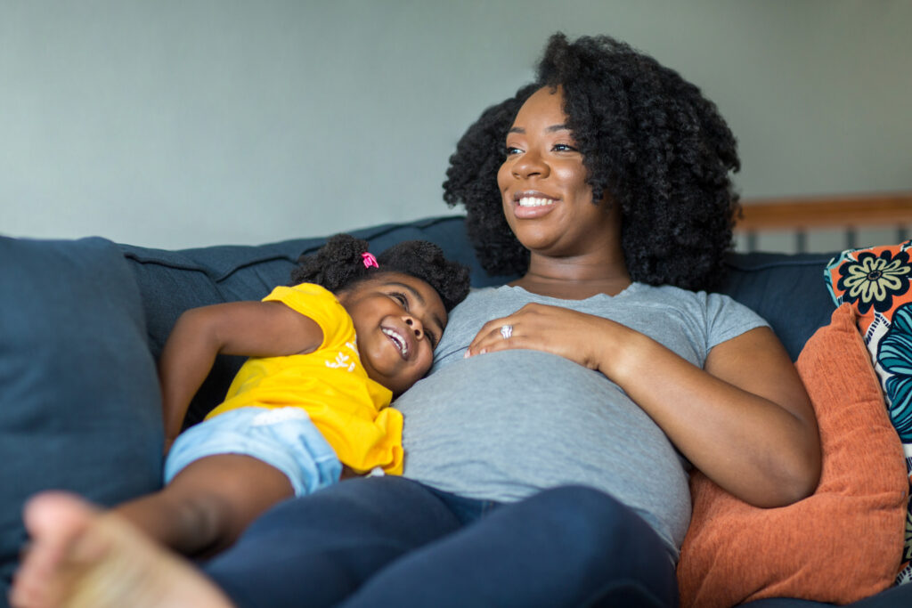 Pregnant Mom and Toddler Daughter sharing a laugh while snuggled on a sofa 